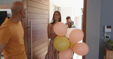 Smiling woman leading friends arriving with pastel balloons greeting host at front door
