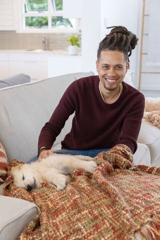 Smiling man with dreadlocks petting sleeping golden retriever puppy on cozy couch