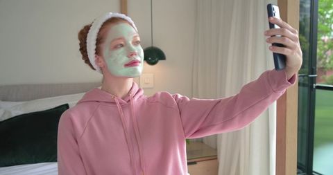 Young woman taking skincare selfie with green face mask in cozy bedroom natural light