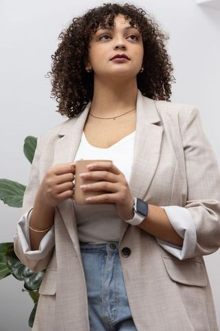 African American woman holding tan mug wearing beige blazer looking contemplative against pale wall