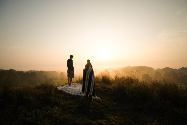 Family standing on grassy hill covered with blanket, enjoying sunrise over scenic countryside. Misty morning with fog creating tranquil atmosphere. Ideal for concepts of bonding, peace, nature, and serene moments during golden hour.