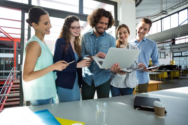 Group of diverse business professionals smiling and collaborating over a laptop in a modern office environment. Ideal for use in articles or advertisements related to teamwork, business meetings, office culture, and collaborative workspaces.