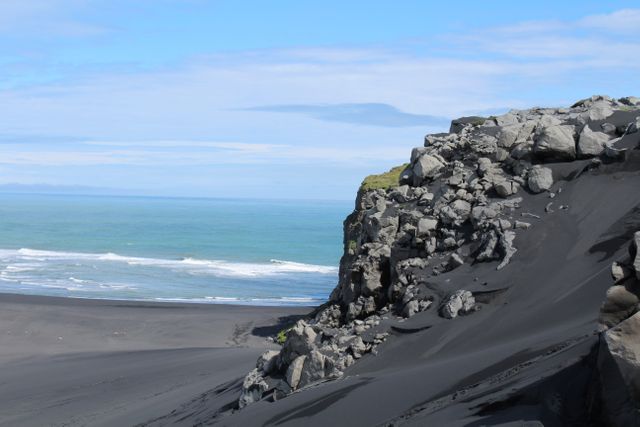 This breathtaking scene captures the stark contrast of black sand against the blue ocean at a secluded black sand beach, complete with a rugged rocky cliff and gentle waves lapping at the shore under a clear blue sky. Ideal for use in travel blogs, inspirational nature prints, coastal tourism advertisements, or geological study materials.