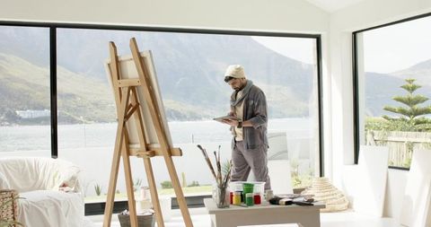 Artist working on tablet next to easel in sunlit lakeside studio with mountain view