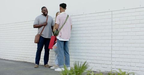 Diverse young friends chatting laughing leaning on white brick wall with red shopping bag, casual st