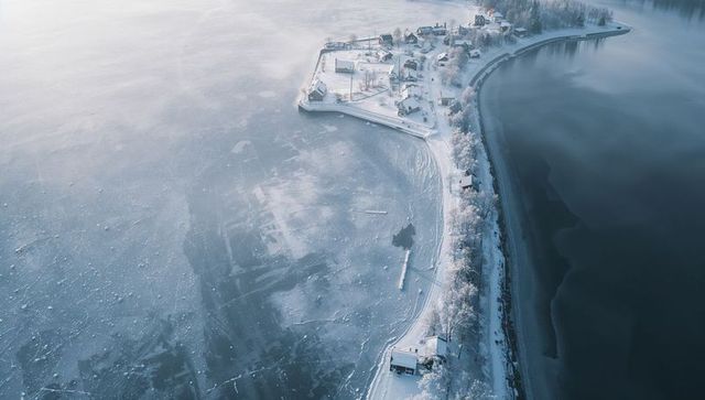 Aerial winter peninsula curving into frozen lake with snow-covered cabins and coastal road