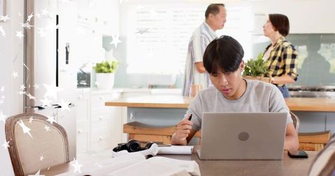 Man working remotely on laptop at sunlit kitchen table with papers, headphones and phone