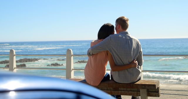 Two individuals are enjoying a romantic moment as they sit and embrace on a seaside bench, overlooking the ocean. Perfect for use in advertisements and campaigns focused on love, relationships, travel, and leisure. This image can also complement content promoting seaside destinations, summer vacations, and outdoor activities.