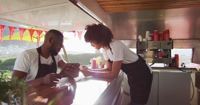 Two food truck workers in black aprons, a man and a woman, are discussing an order on a tablet. Ideal for representing small business operations, teamwork, outdoor restaurant services, technological integration in food industry, and customer engagement.
