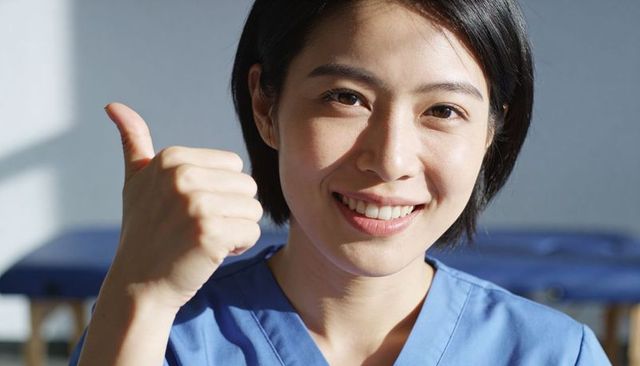 Smiling asian nurse giving thumbs-up in blue scrubs near clinic exam table, sunlit closeup