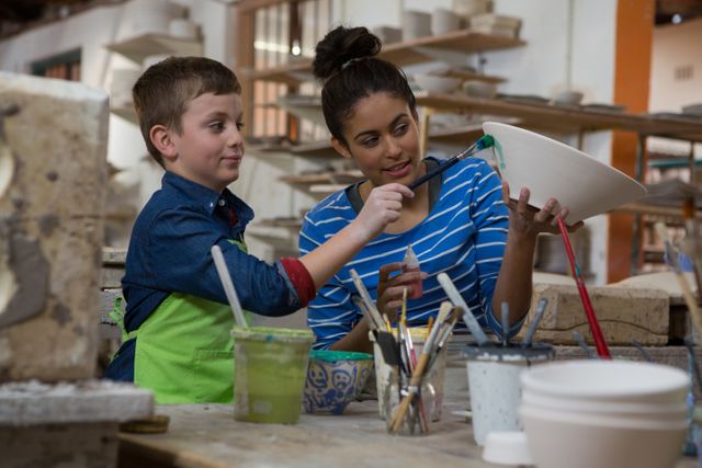 Female potter assisting her son in painting a bowl at a pottery shop. They are surrounded by pottery tools and materials, indicating a creative and educational environment. This image can be used for promoting family activities, art classes, creative workshops, and educational programs.