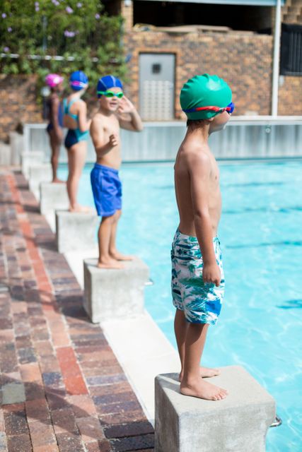 Children standing on start blocks at poolside, wearing swim caps and goggles, preparing for a swim race. Ideal for use in content related to children's sports, swimming competitions, summer activities, teamwork, and outdoor fun.