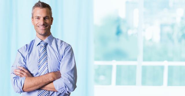 Confident businessman standing with arms crossed near large glass window, smiling, appearing professional and approachable. Ideal for business-themed content, corporate websites, leadership articles, success stories, or advertisements featuring modern office environments.