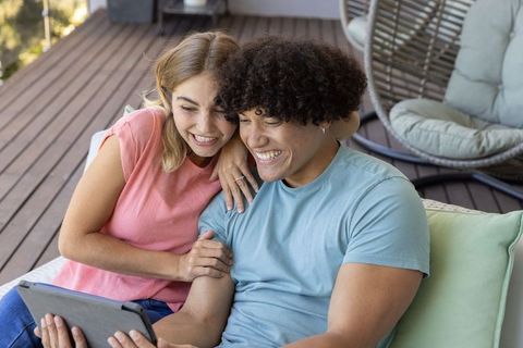Young couple cuddling on sunlit patio sofa while watching tablet, cozy outdoor romance