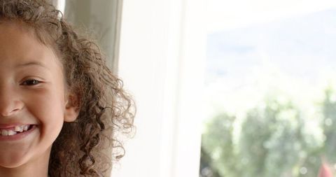 Smiling girl with curly hair by sunlit window, closeup portrait conveying childhood joy