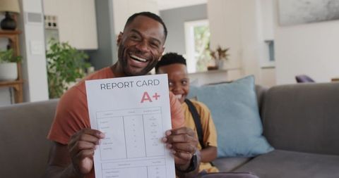 African-american father celebrating teen son a+ report card on home couch with big smile
