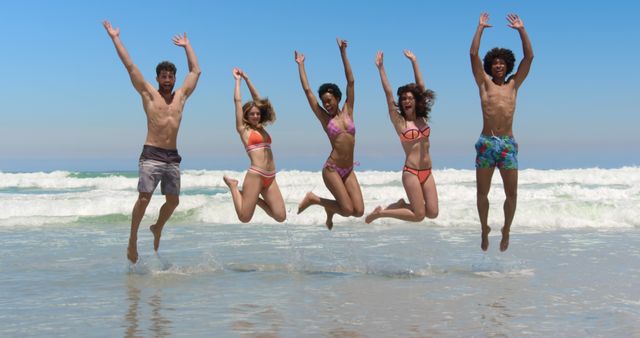 Group of happy friends jumping while standing in shallow water at the beach, enjoying summer together. Ideal for promotions related to summer holidays, beachwear, group activities, and travel destinations highlighting friendship and outdoor fun.