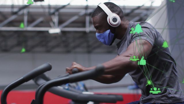 A man exercising on a stationary bike, wearing headphones and a protective mask in a gym setting. Overlay of digital network icons represents virtual connections and technology advancement. This image can illustrate topics on fitness technology, digital fitness apps, and hybrid workout environments combining virtual and physical worlds.