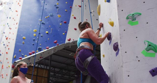 A woman is engaging in rock climbing at an indoor climbing gym, scaling the artificial wall while secured with a harness. An instructor or teammate is present wearing a face mask, ensuring safety and providing guidance. This visual can be used for promoting fitness activities, teamwork, training programs, or climbing gyms.