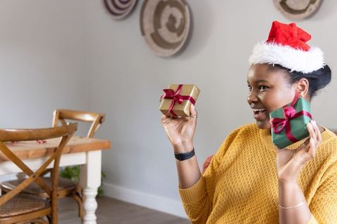 African american woman wearing santa hat holding two wrapped christmas gifts in cozy dining room