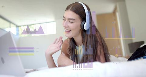 Waving young woman wearing headphones using laptop on bed with floating data visualizations