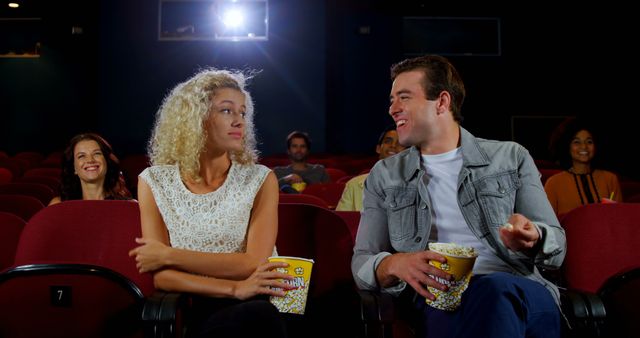 Couple sitting and chatting in a movie theater while eating popcorn. They seem to be on a date and enjoying each other's company. Audience members are visible in the background, suggesting a lively and shared entertainment experience. Use for dating services, social interactions, leisure activities, or movie-related promotions.