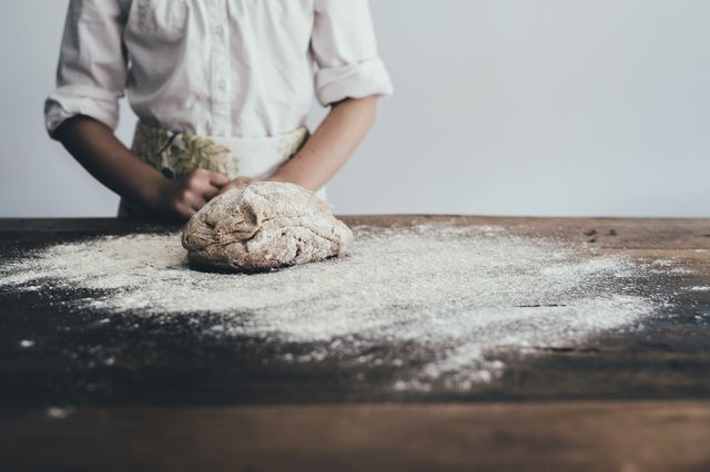 Person kneading dough on a floured wooden table. The person is wearing a white shirt, with part of an apron visible. Ideal for blogs or articles about baking, culinary arts, homemade recipes, rustic kitchen aesthetics, or cooking demonstrations.