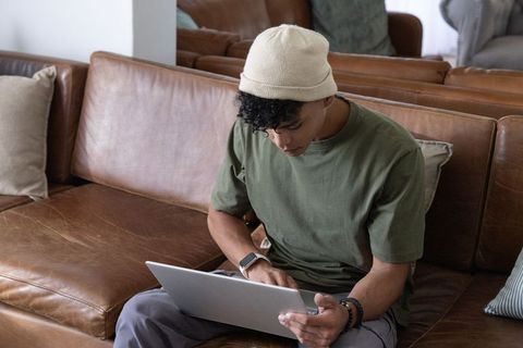 Young african american man working on laptop on leather sofa in cozy living room