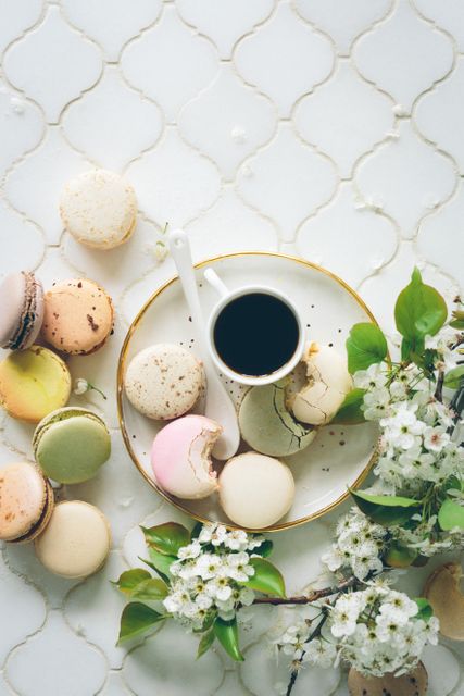 An assortment of colorful macarons arranged around a cup of black coffee on a white table. Blossoming branches add a touch of springtime elegance to the scene. Ideal for use in culinary blogs, gourmet café promotions, elegant dessert presentations, or spring-themed event invitations.