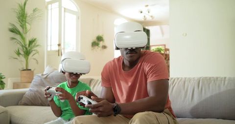 African American Father and Son Wearing VR Headsets Playing Video Games on Couch at Home