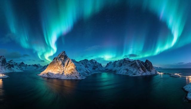 Aurora arching over lit snowy peak and reflecting in calm fjord under arctic night sky