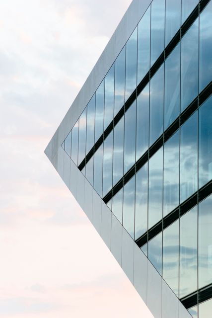 An artistic angle of a contemporary glass building showcasing its geometric design and reflecting the sky. Ideal for use in architectural magazines, urban planning presentations, construction industry materials, or as a background for corporate and real estate visuals.
