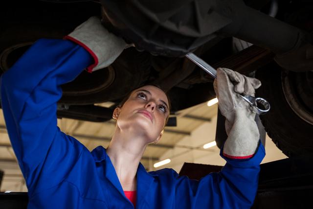 Female mechanic wearing blue uniform and gloves using a wrench to service a car in a professional repair garage. Perfect for illustrating topics related to gender equality in trades, promoting automotive repair services, or showcasing mechanical expertise.