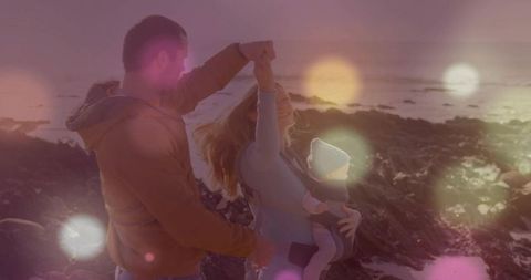 Twirling parents with baby in carrier on rocky shore at sunset golden hour family joy