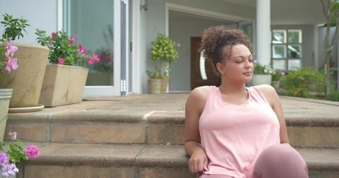 African american woman relaxing on sunlit porch steps in pink athleisure with planters