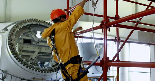 Construction worker wearing safety harness working on scaffold in industrial setting. Image is ideal for articles, blogs, or presentations related to construction safety, occupational health, industrial work, or engineering projects. Could also be used in training materials, company brochures, or safety equipment promotions.