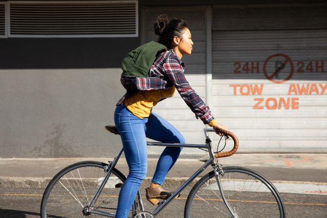 Side view of a biracial woman with long dark hair in a hair bun out and about in the city streets during the day, riding her bicycle in the sun wearing a backpack, a grey building in the background.
