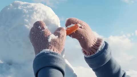 Placing carrot nose on snowman head with pink mittens and blue cuffs under clear winter sky