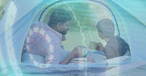 Father and son camping inside tent sharing hot drinks while bonding outdoors