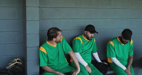 Baseball teammates sitting on dugout bench resting in green uniforms