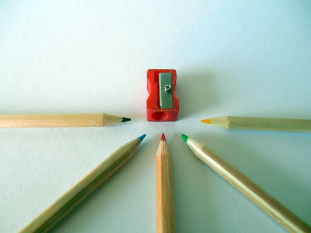 Five colored pencils are arranged in a circle around a red sharpener on a white background. This image can be used for educational materials, art and craft supplies advertisements, back-to-school promotions, or creative inspiration content for students and artists.
