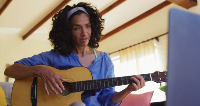 Biracial woman playing guitar while sitting on the couch in front of laptop at home. staying at home in self isolation in quarantine lockdown