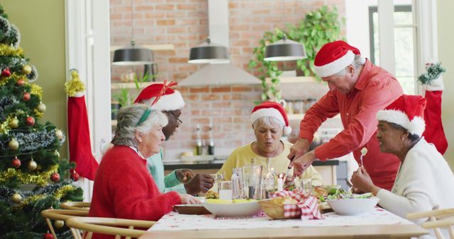 Senior friends sharing a meal around a table, wearing Santa hats in a decorated kitchen. Ideal for use in holiday greeting cards, advertisements for senior community activities, or festive cookbooks showcasing holiday traditions.