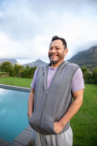 Smiling mature asian man in gray hoodie beside pool with mountain and garden backdrop