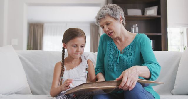 Grandmother and granddaughter are sitting on a sofa looking through a family photo album. The elderly woman is pointing at pictures while engaging the young girl in conversation. Ideal for themes of family bonding, passing down traditions, childhood memories, and intergenerational connections.