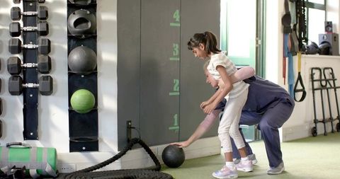 Female fitness coach guiding young girl reaching medicine ball at wall height markers