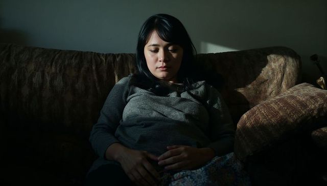 Asian woman sitting on sofa looking down in moody windowlight, contemplative solitude