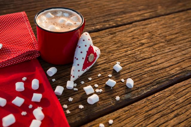 Hot chocolate with marshmallows in a red cup, accompanied by a heart-shaped decoration and a red polka dot napkin on a wooden table. Ideal for holiday-themed promotions, Christmas greeting cards, cozy winter scenes, and festive social media posts.