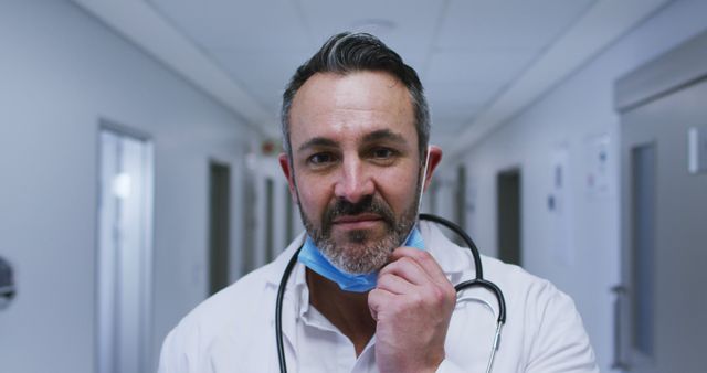 Male doctor, wearing a white coat, adjusts face mask while standing in a hospital corridor. The doctor looks confident and focused, capturing the essence of dedication in healthcare. Ideal for editorial content, healthcare advertisements, medical brochures, and websites emphasizing health services and professional care.