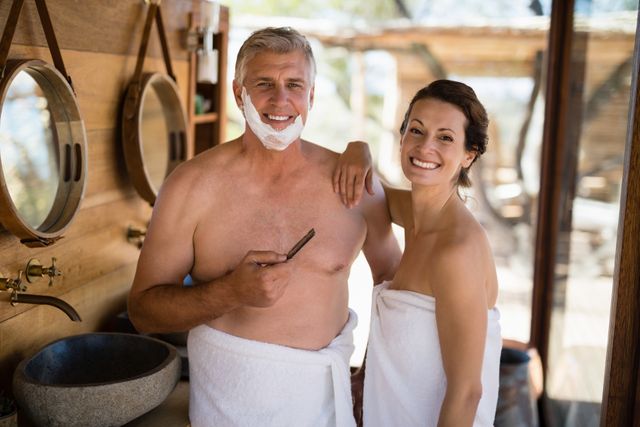 Couple enjoying a relaxing moment while shaving in a rustic cottage during their safari vacation. Both are wrapped in towels, smiling, and standing by a bathroom sink with mirrors. Ideal for use in travel brochures, lifestyle blogs, and advertisements promoting vacation experiences, relaxation, and couple bonding.